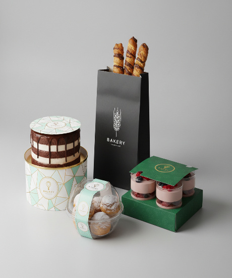 Assorted gourmet bakery items including a layered chocolate cake in a geometric patterned box, pastry sticks in a tall black bag, fruit parfaits in a green carrier, and cream puffs in a clear domed container.