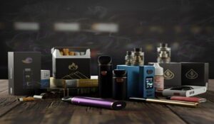 A diverse collection of smoking and vaping products is spread across a rustic wooden table against a dark, smoky background. The assortment includes various electronic vape pens, box mods with digital displays, liquid bottles, and several styles of traditional cigarette packs.