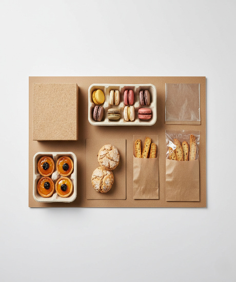 An overhead shot shows various baked goods in eco-friendly brown paper and molded pulp packaging on a tan surface.
